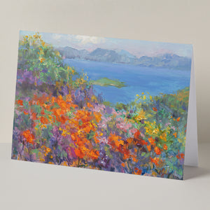 Fine art greeting card displayed on background, including orange California poppies and other wildflowers, hills, lake, based on an original landscape oil painting by Oksana Johnson. Yellow, orange, and purple flowers, green grass, blue water, white clouds.