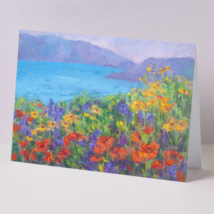 Fine art greeting card displayed on background, including orange California poppies and other wildflowers, hills, lake, based on an original landscape oil painting by Oksana Johnson. Yellow, orange, and purple flowers, green grass, blue water, white clouds.