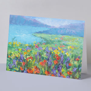 Fine art greeting card displayed on background, including orange California poppies and other wildflowers, hills, green meadow, lake, based on original landscape oil painting by Oksana Johnson. Yellow, orange, and purple flowers, green grass, blue water, white clouds.