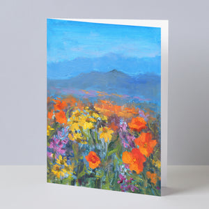 Fine art greeting card displayed on background, including orange California poppies and other wildflowers, hills, sky,  based on an original landscape oil painting by Oksana Johnson. Yellow, orange, and purple flowers, green grass, blue hills and sky.