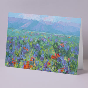 Fine art greeting card displayed on background, including orange California poppies and other wildflowers, hills, lake, based on an original landscape painting by Oksana Johnson. Orange, and purple flowers, green grass, blue water, white clouds, hills in background.